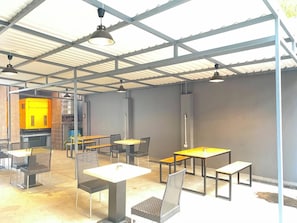 An indoor dining area with tables and chairs, a high ceiling with metal beams, and a kitchen area in the background.
