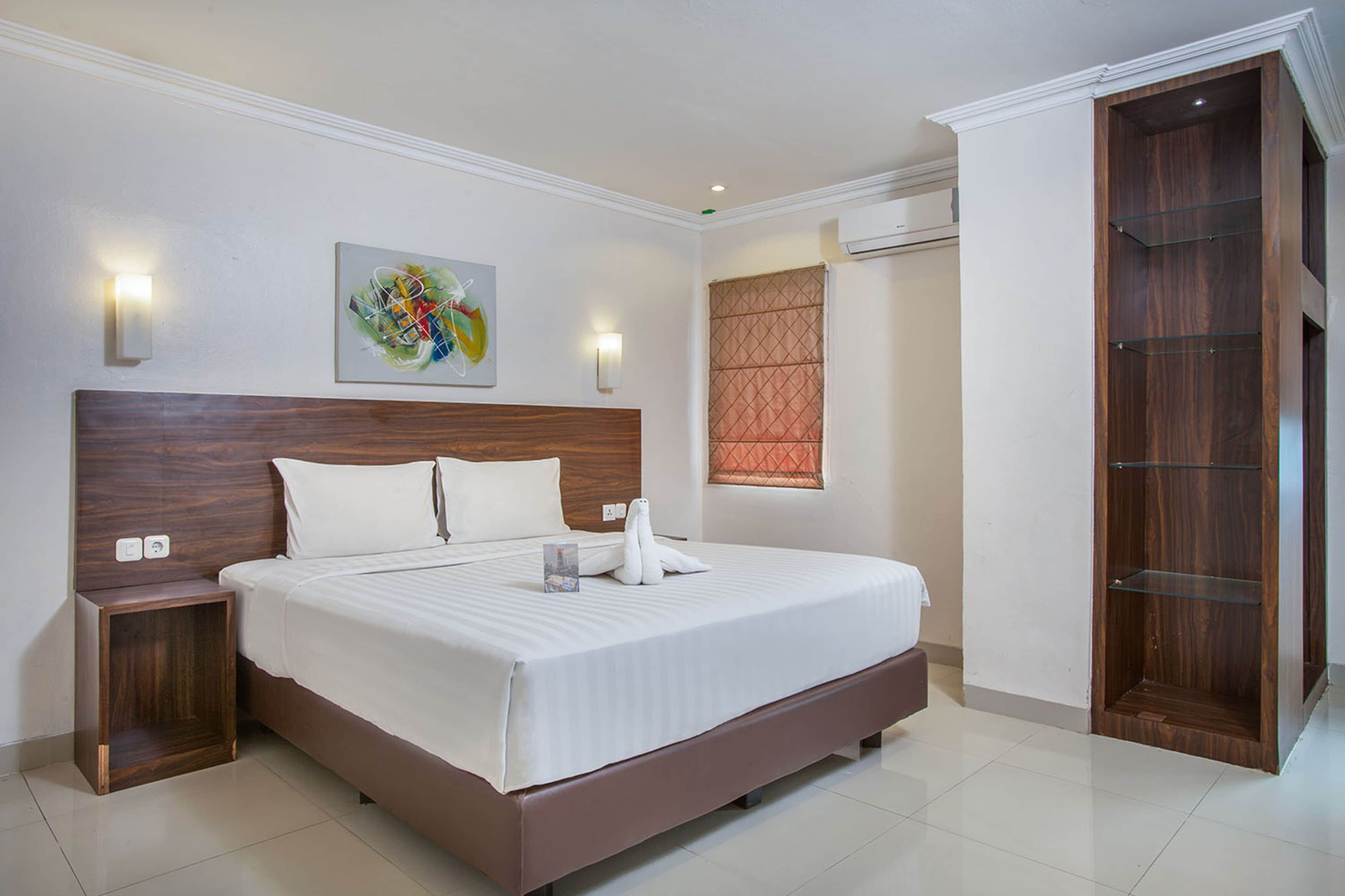 A modern hotel room with a large bed, a wooden headboard, a nightstand, a wall-mounted light, a window with blinds, and a wooden wardrobe.