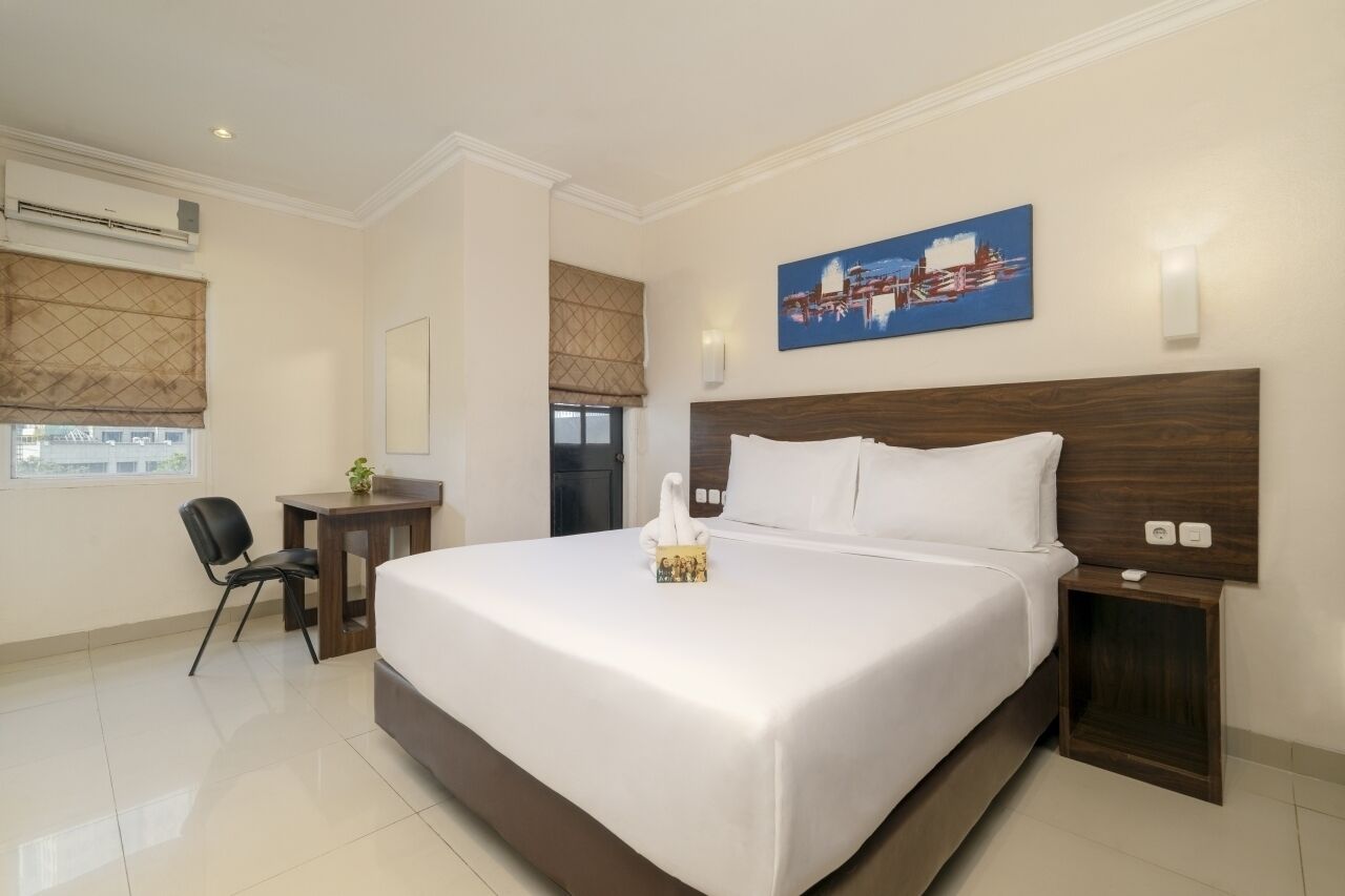 Habitación de hotel con una cama grande, un escritorio y una silla. Hay un cuadro en la pared y un equipo de aire acondicionado sobre la cama.