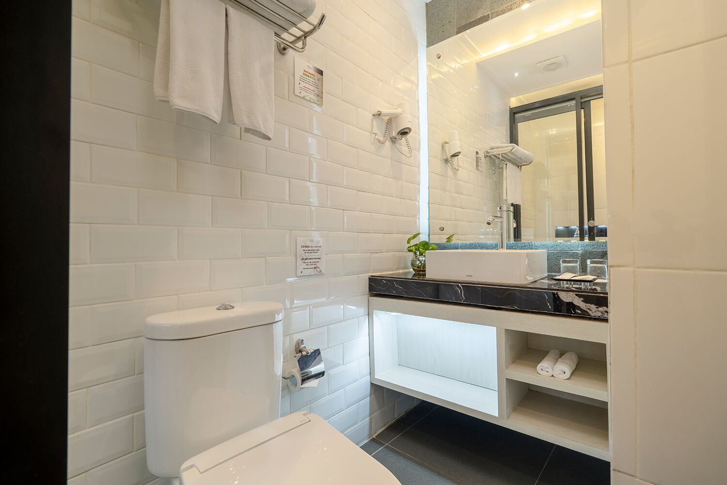 Une salle de bain moderne avec des carreaux blancs, un comptoir en marbre noir, des toilettes, un lavabo avec un miroir et une douche.