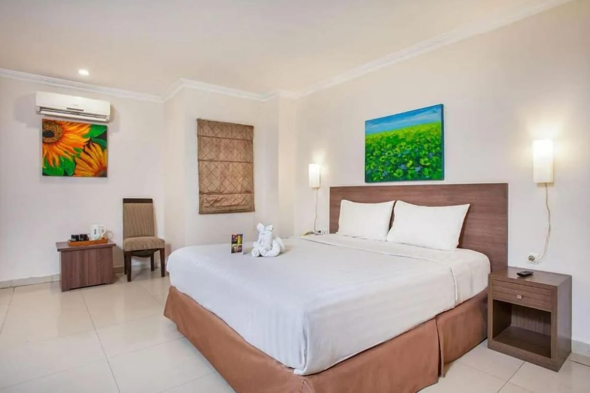 A hotel room with a large bed, a chair, a nightstand, an air conditioning unit, and two wall-mounted lamps.