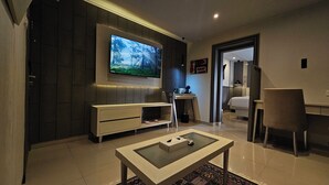 A modern living room with a flat-screen TV mounted on the wall, a white sofa, a glass-top coffee table, and a small desk with a chair. - 1O1 URBAN Jakarta Thamrin (Jakarta)