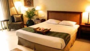 A neatly arranged hotel room with a large bed, a bedside table with a lamp, a chair, and a potted plant. - The Graha Cakra (MALANG)