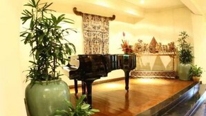 A grand piano in a well-lit room with a wooden floor, surrounded by large green plants in vases and a decorative wall hanging. - The Graha Cakra (MALANG)