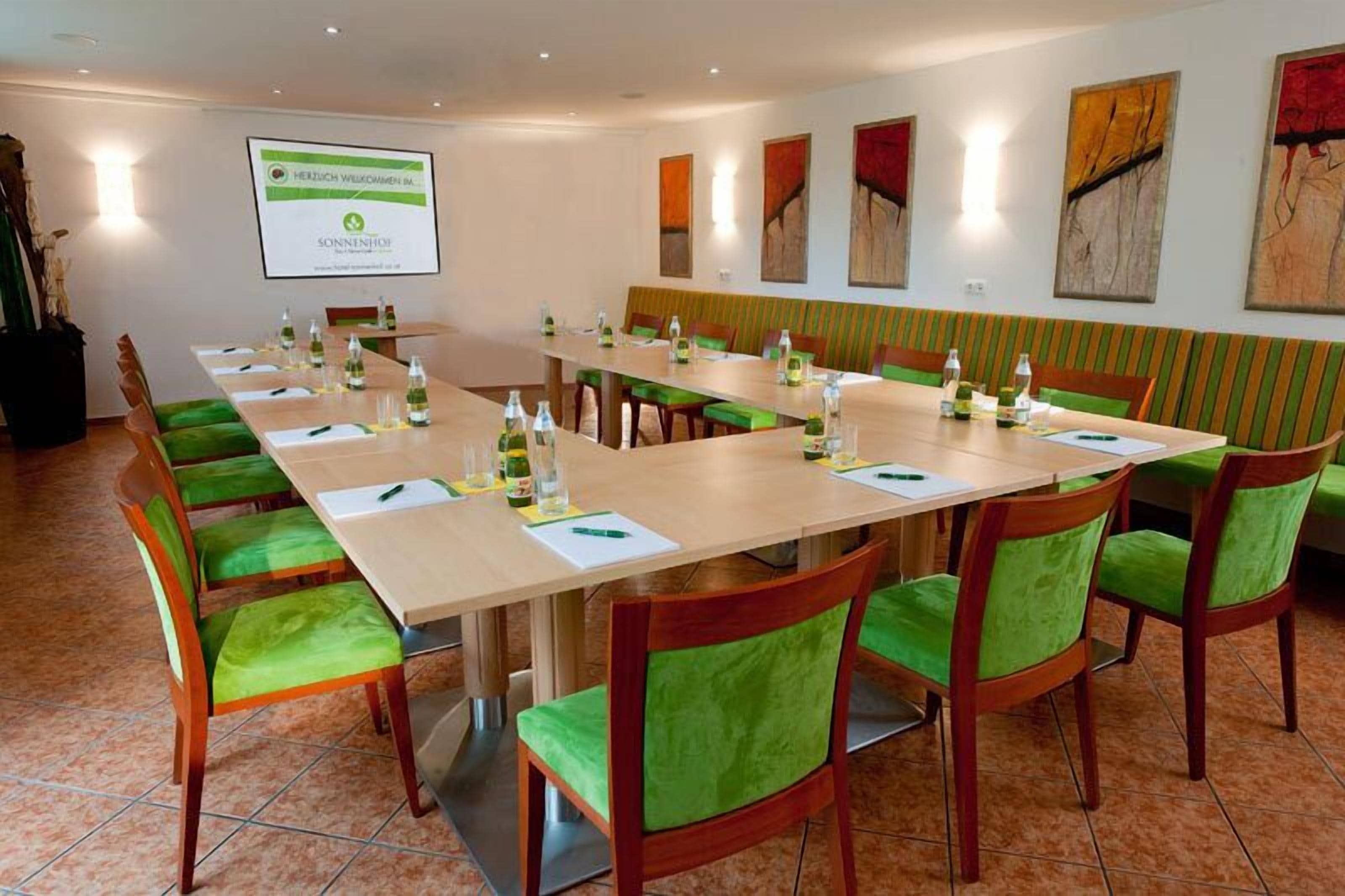 A conference room with a long table, green cushioned chairs, and a projector screen on the wall.
