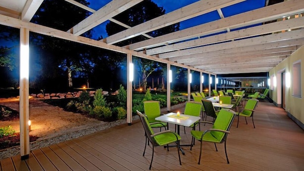 Un patio al aire libre con vigas de madera, sillas y mesas verdes, rodeado de jardines y árboles.