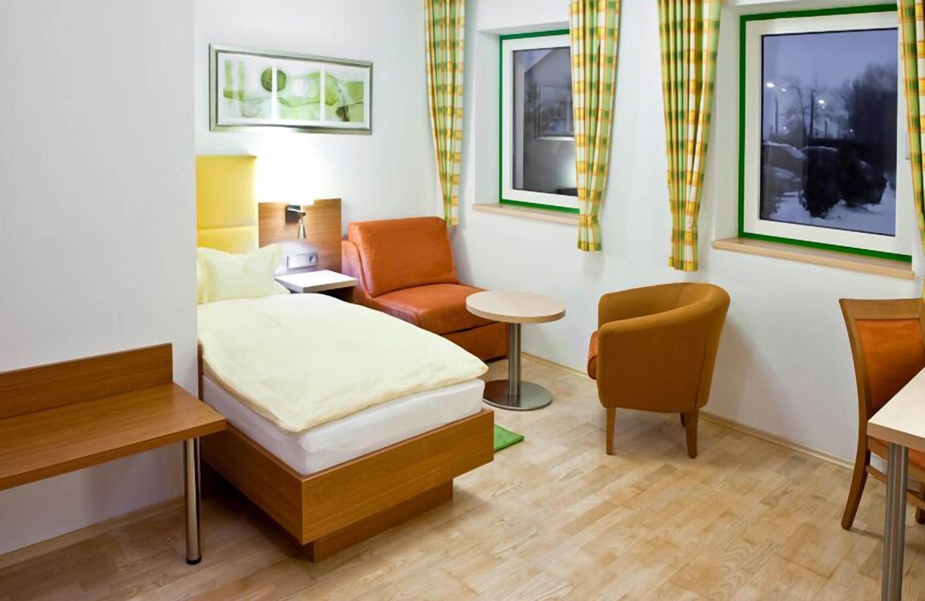 Una camera d'albergo compatta con un letto singolo, una scrivania, una sedia e un tavolino rotondo.