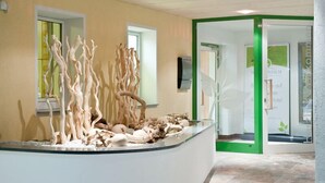 Un área de recepción con un mostrador central de vidrio que exhibe esculturas abstractas. Hay dos ventanas a la izquierda y una puerta con marco verde a la derecha.