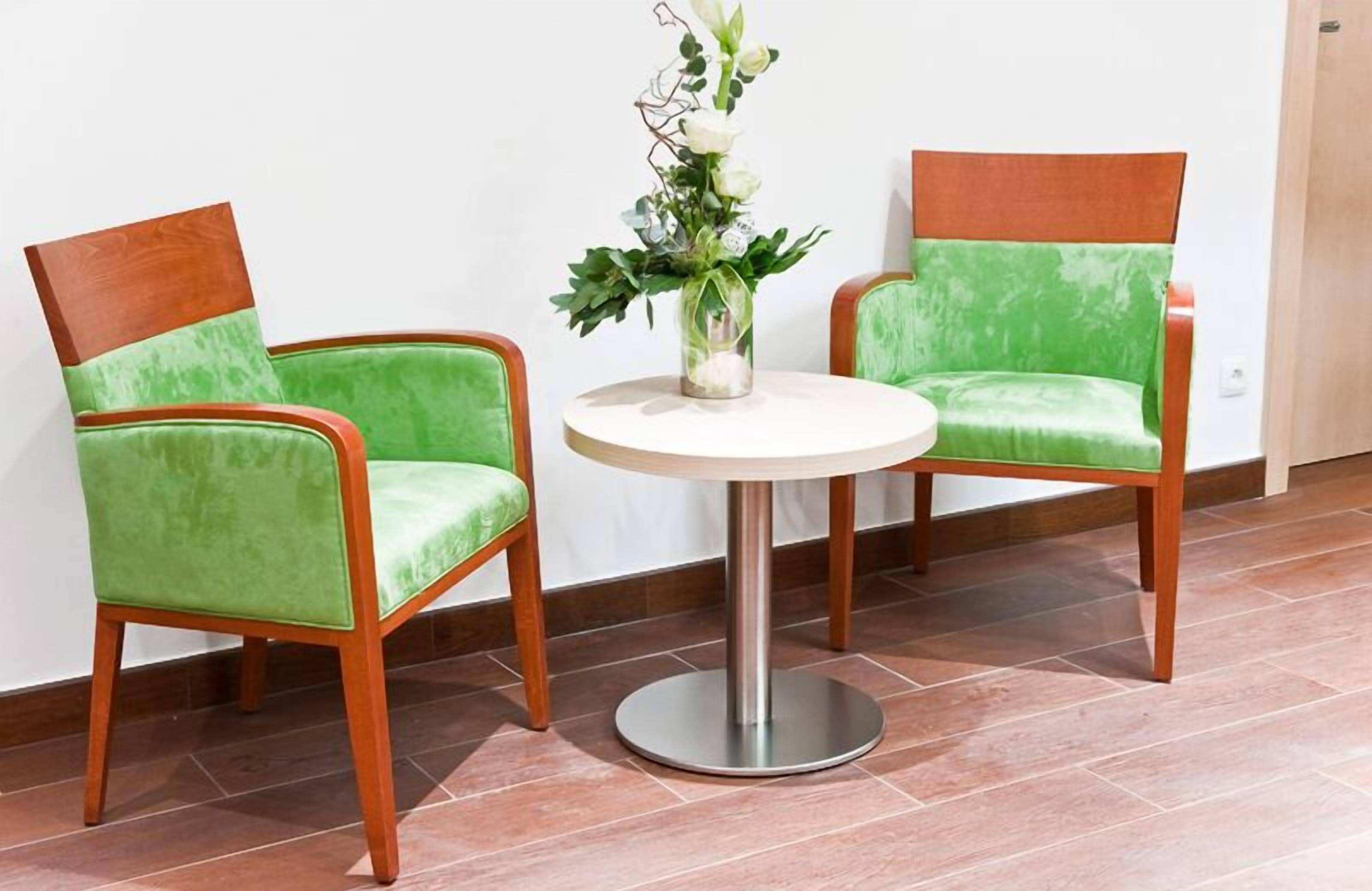 Dos sillones verdes tapizados con armazón de madera y una mesa blanca con base metálica, ubicados frente a una pared blanca con piso de baldosas.
