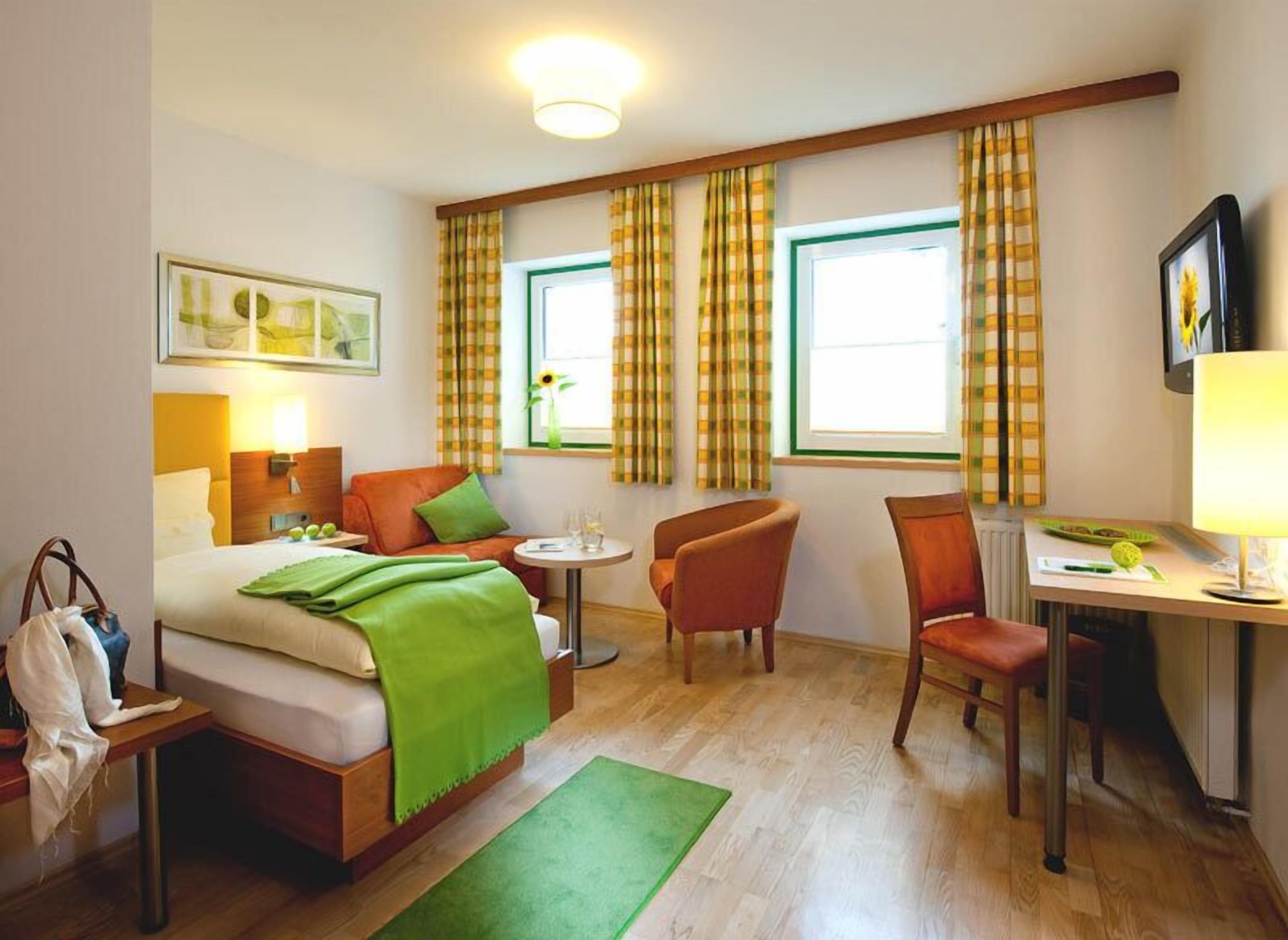A hotel room with a bed, a desk, two chairs, a table, a green rug, and curtains.