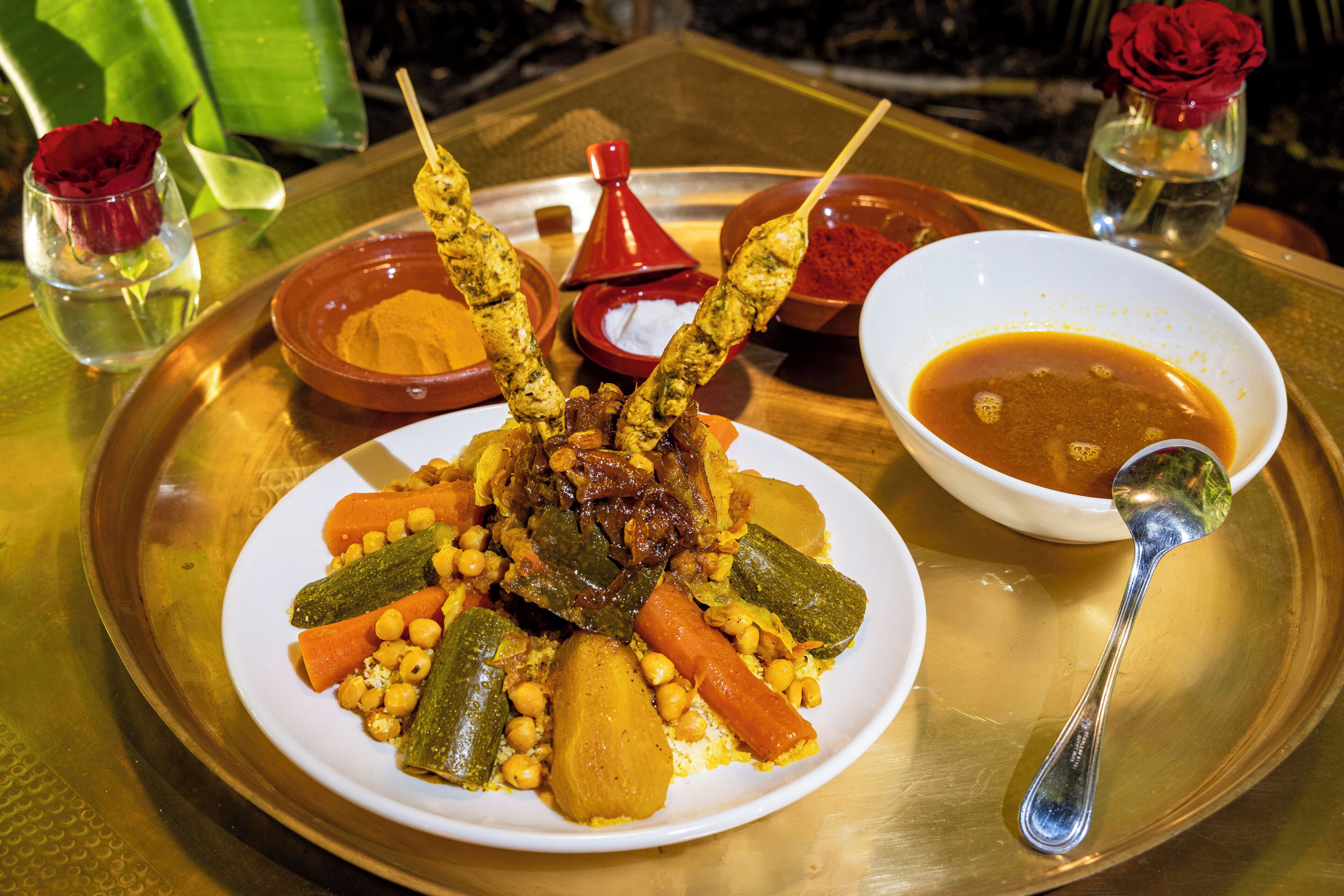 A plate of food with vegetables and skewers, accompanied by bowls of sauces and a spoon on a tray.