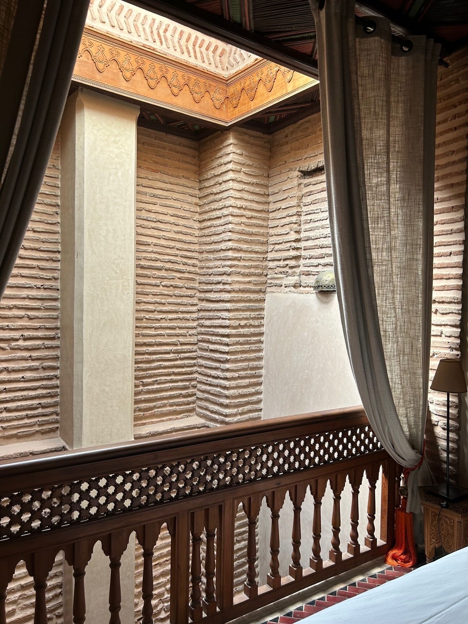 Ein Zimmer mit Blick auf einen Innenhof, ausgestattet mit einem hölzernen Balkon mit aufwendiger Geländerkonstruktion, einem Bett mit weißer Bettdecke und einer Lampe auf einem Nachttisch.