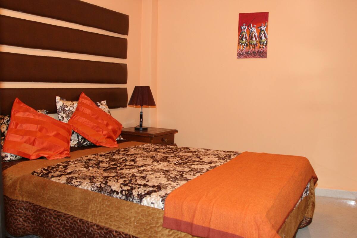 Una cama bien hecha con una colcha estampada y dos almohadas naranjas, una lámpara de noche y un cuadro en la pared.