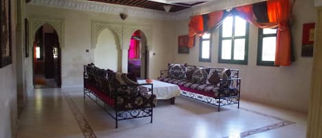 A spacious room with a high ceiling, ornate ceiling design, and a large window with orange curtains. There are two sofas and a small table in the center.