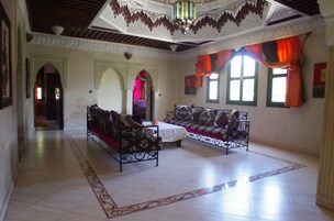Una habitación amplia con un techo alto, un diseño ornamental en el techo y un gran ventanal con cortinas naranjas. Hay dos sofás y una mesita en el centro.