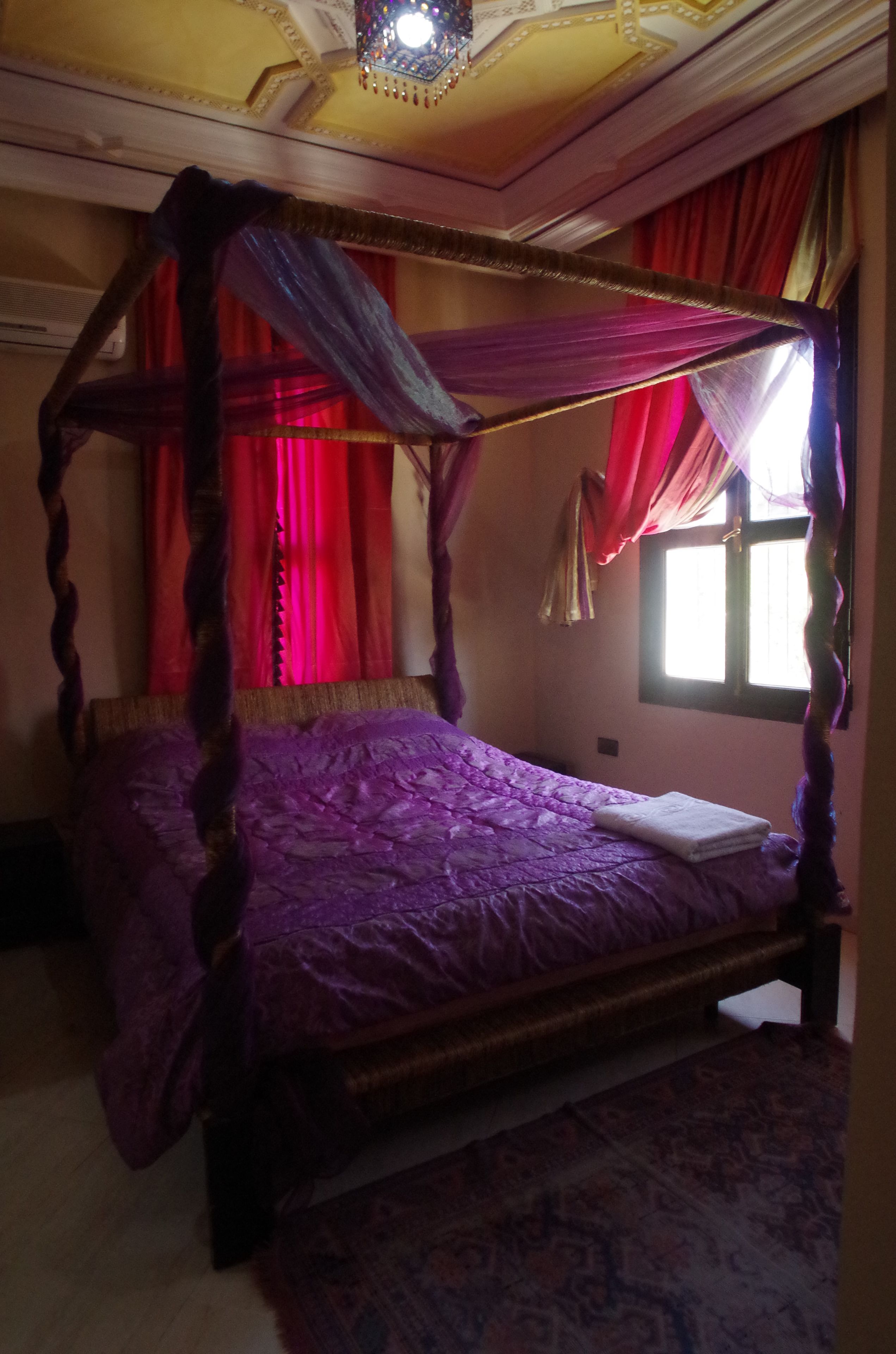 A four-poster bed with a purple bedspread in a room with red curtains and a patterned carpet.