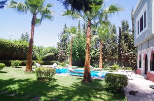 Zona de piscina con sillones de descanso, palmeras y un edificio de fachada roja y blanca.