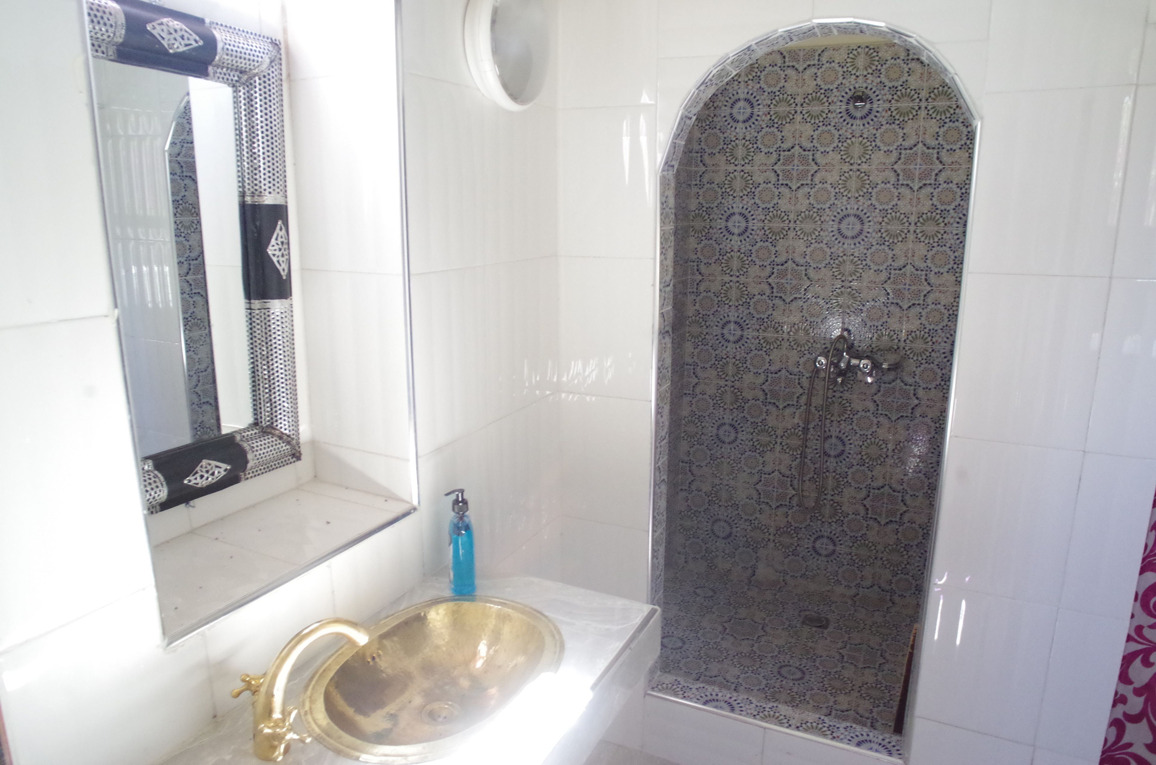 Un baño con pared de azulejos blancos, un lavamanos dorado, un dispensador de jabón azul y una ducha con baldosas estampadas.
