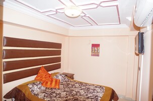 Un dormitorio con cabecera, cama con ropa de cama estampada, mesita de noche pequeña, televisor montado en la pared y lámpara en el techo.