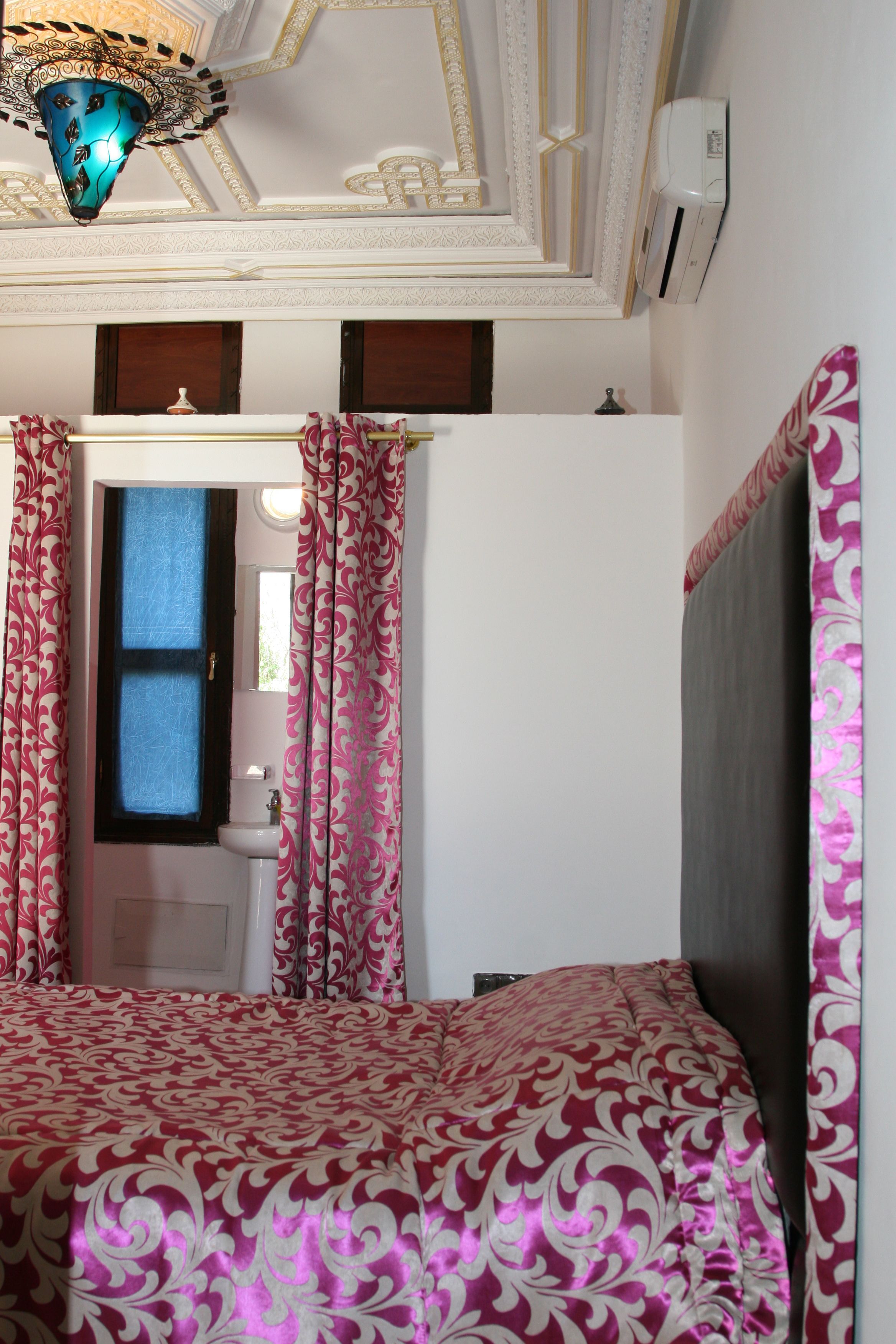 Une chambre à coucher avec un lit, des rideaux, un luminaire au plafond et une salle de bain visible à travers une porte.