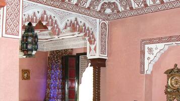 Une pièce de style traditionnel, ornée de motifs complexes sur les murs et le plafond, avec une grande horloge décorative et un coin salon agrémenté de coussins et d’une petite table.