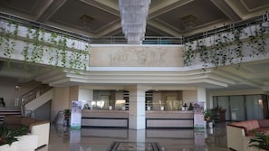 Una hall d'albergo con una reception centrale, un lampadario e piante pensili.
