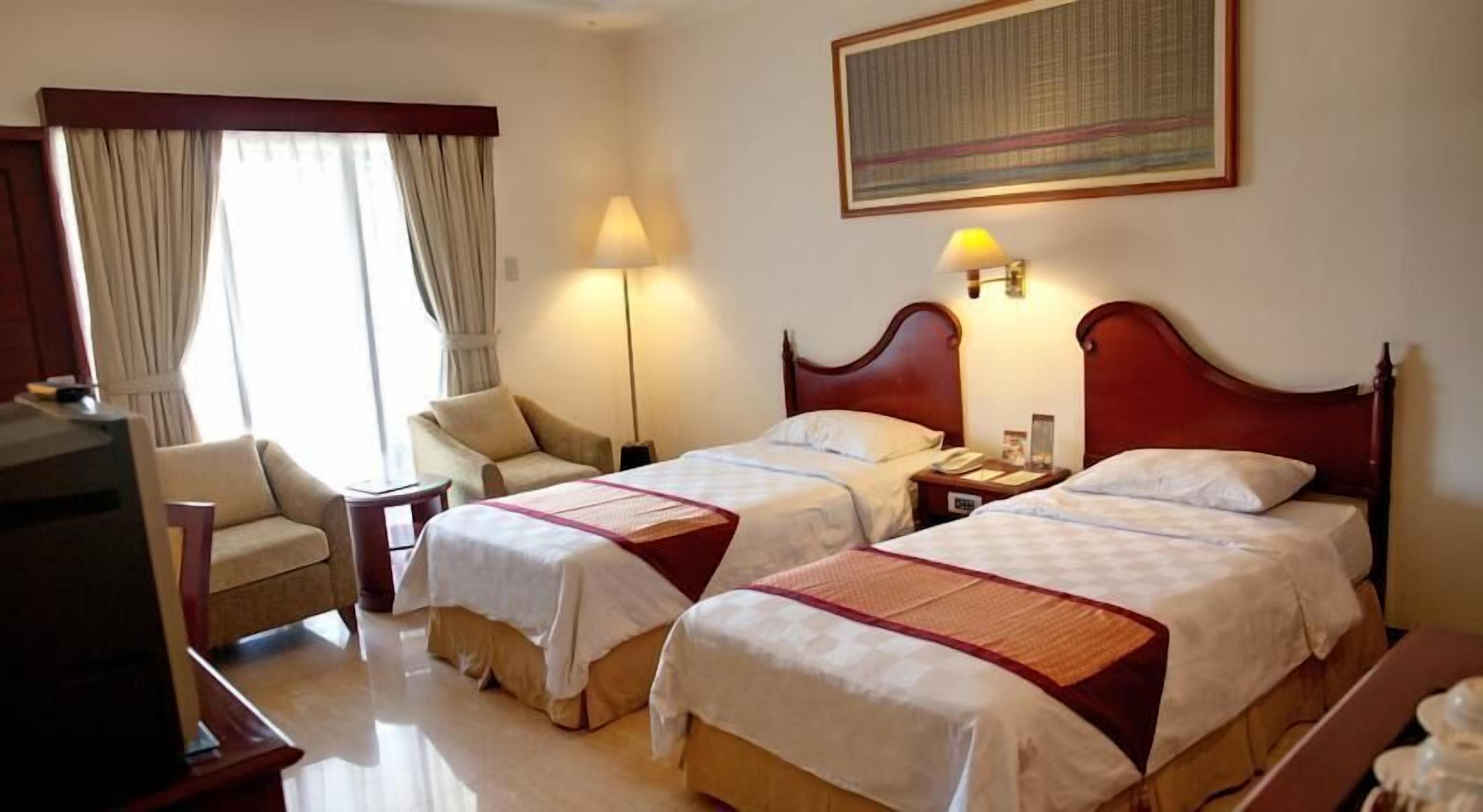 A hotel room with two beds, a desk, a chair, and a lamp.