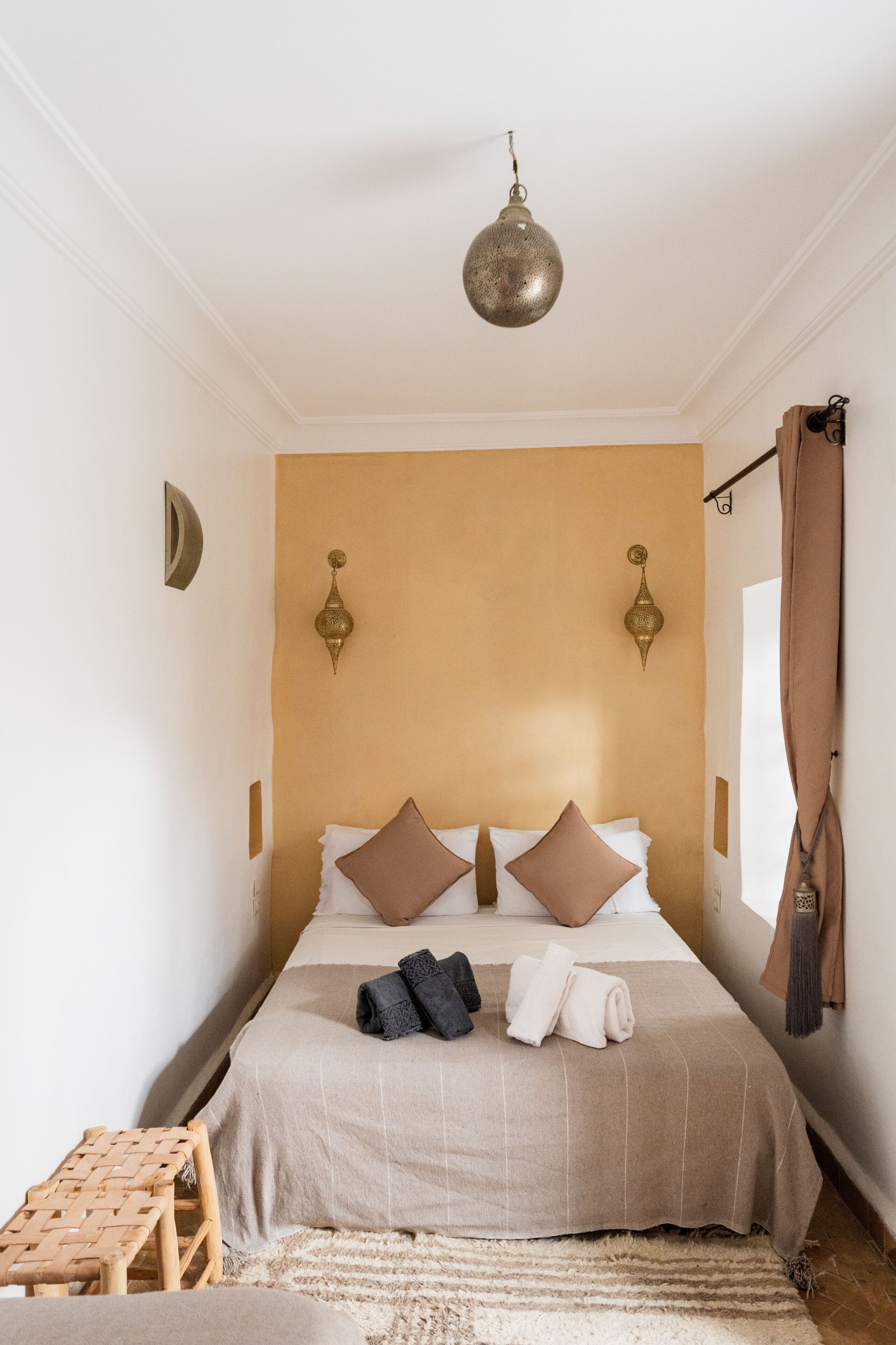 Un dormitorio con una cama, dos lámparas de pared, una lámpara colgante, un taburete de madera y una ventana con cortinas.