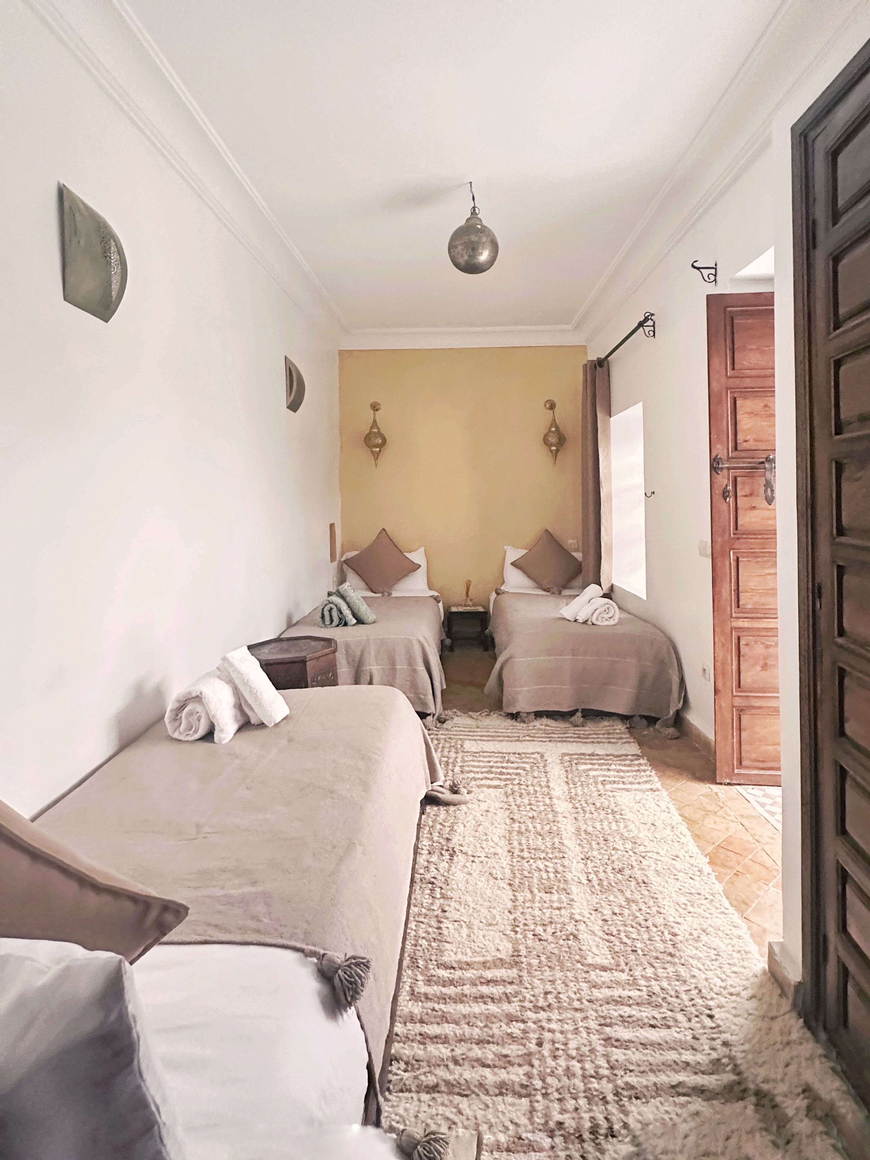 A narrow hallway leading to a bedroom with two beds, a patterned rug, and a wooden door.