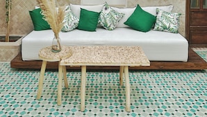Un canapé blanc agrémenté de coussins verts et à motifs, une table en bois dont le plateau est tressé, et un luminaire décoratif suspendu.