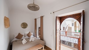Une chambre à coucher avec un lit, un banc, un luminaire suspendu et une vue sur un balcon par une porte ouverte.