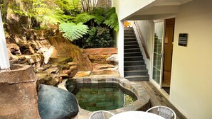 Een stenen hottub met waterval, omringd door rotsen en varens, met een trap die leidt naar een glazen deur.