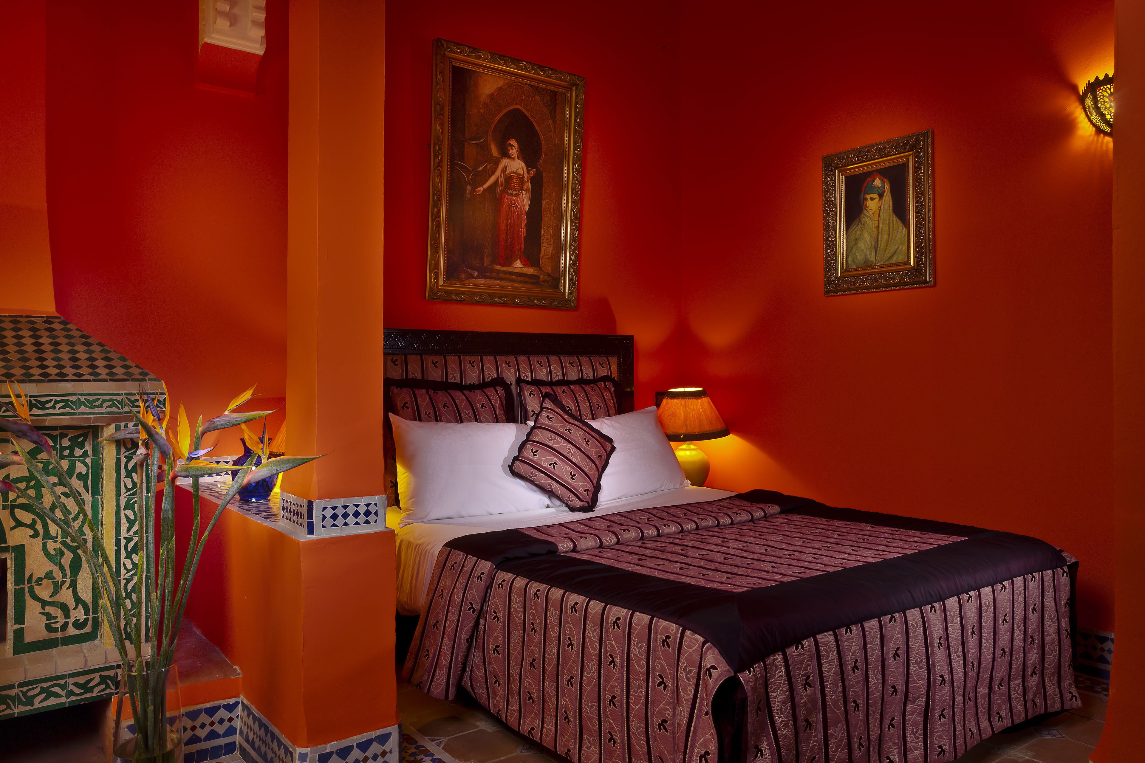 Una habitación con una cama grande, dos cuadros en la pared, una mesita de noche con lámpara y una planta decorativa.