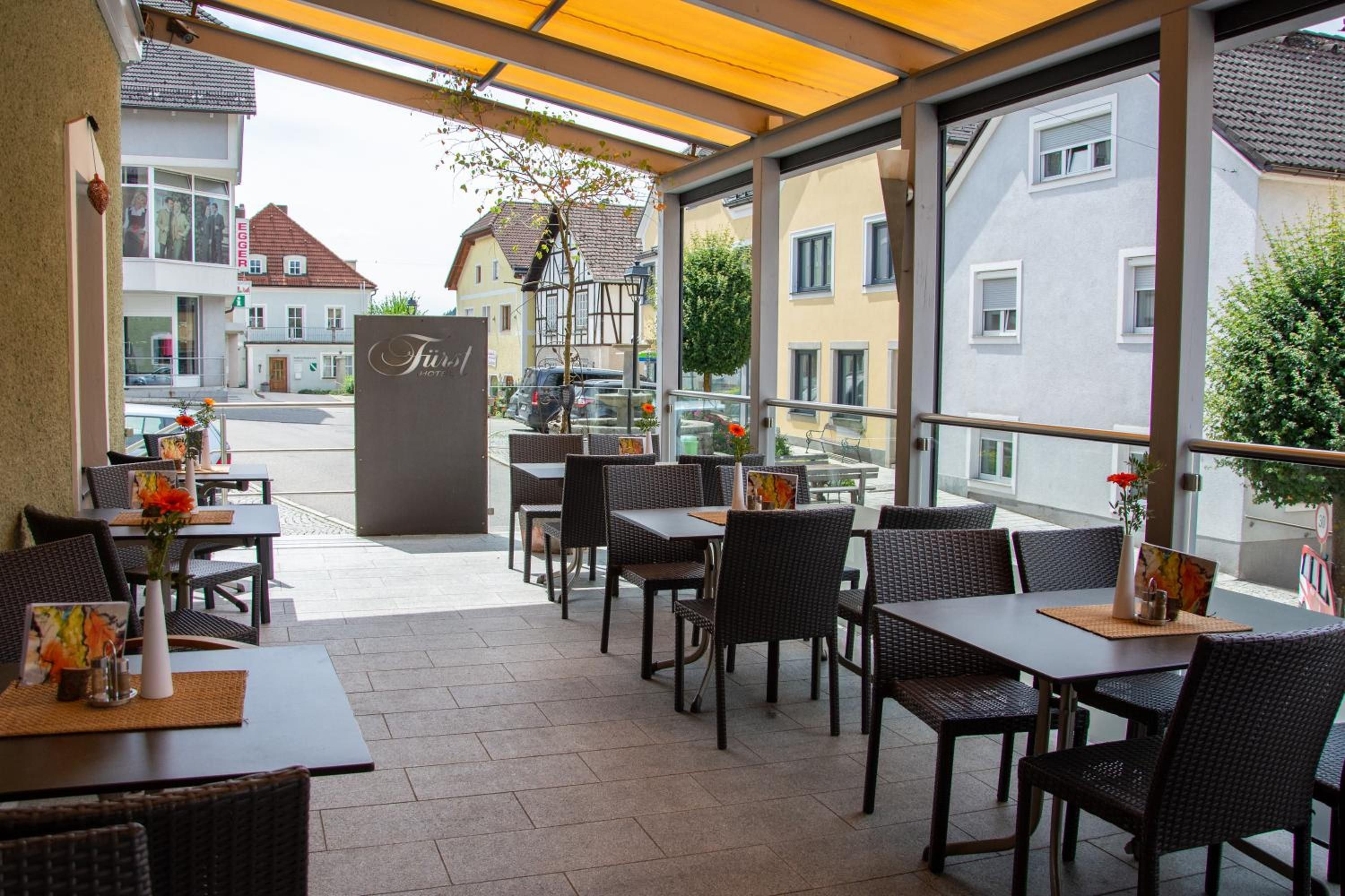 Ein Sitzbereich im Freien mit Tischen und Stühlen, ein Schild mit der Aufschrift 'Friedel Hotel' und Blick auf die Straße und die Gebäude.