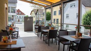 Ein Sitzbereich im Freien mit Tischen und Stühlen, ein Schild mit der Aufschrift 'Friedel Hotel' und Blick auf die Straße und die Gebäude.
