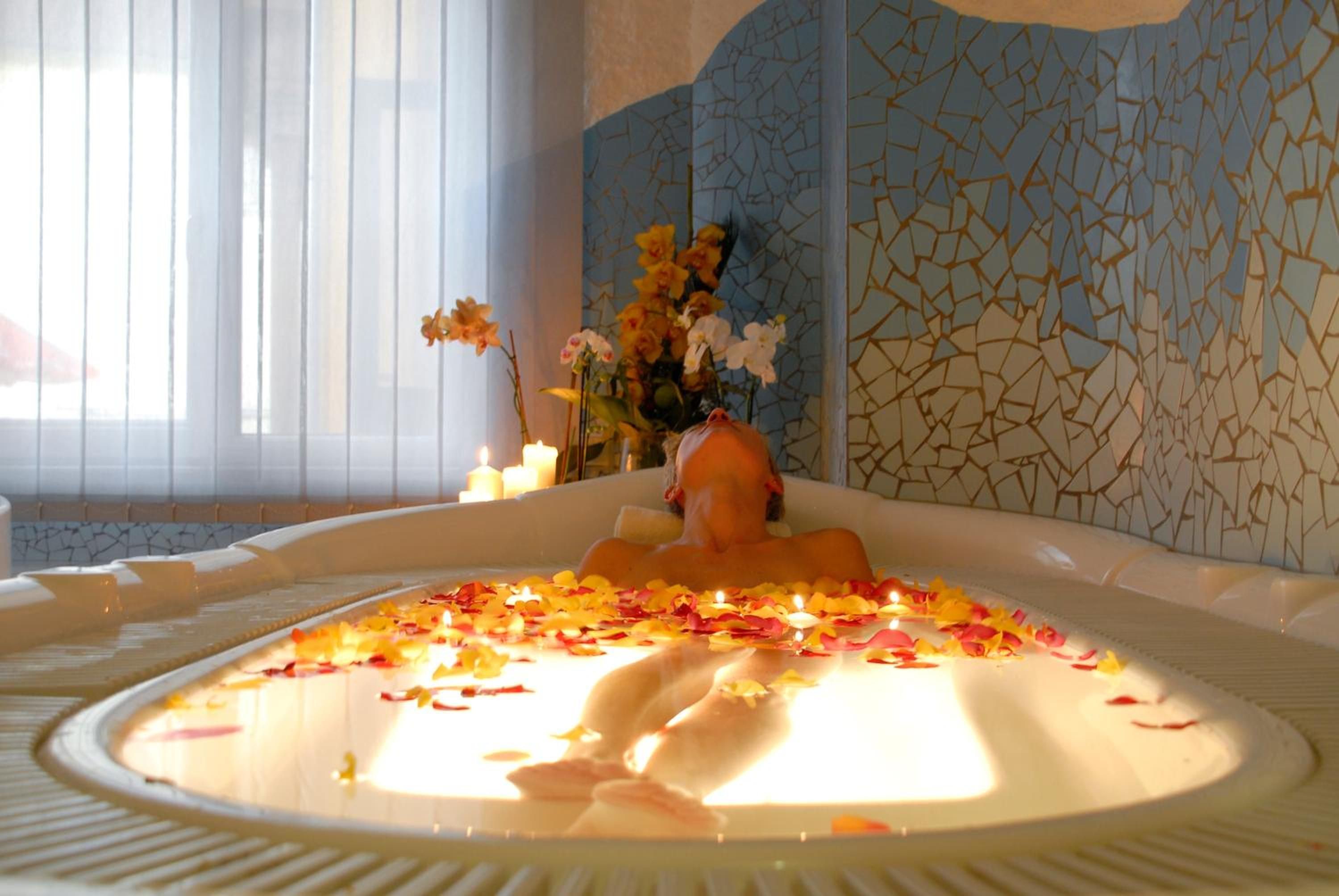 Eine mit Wasser und Rosenblättern gefüllte Badewanne, im Hintergrund eine Blumenvase und Kerzen.