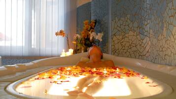 Eine mit Wasser und Rosenblättern gefüllte Badewanne, im Hintergrund eine Blumenvase und Kerzen.
