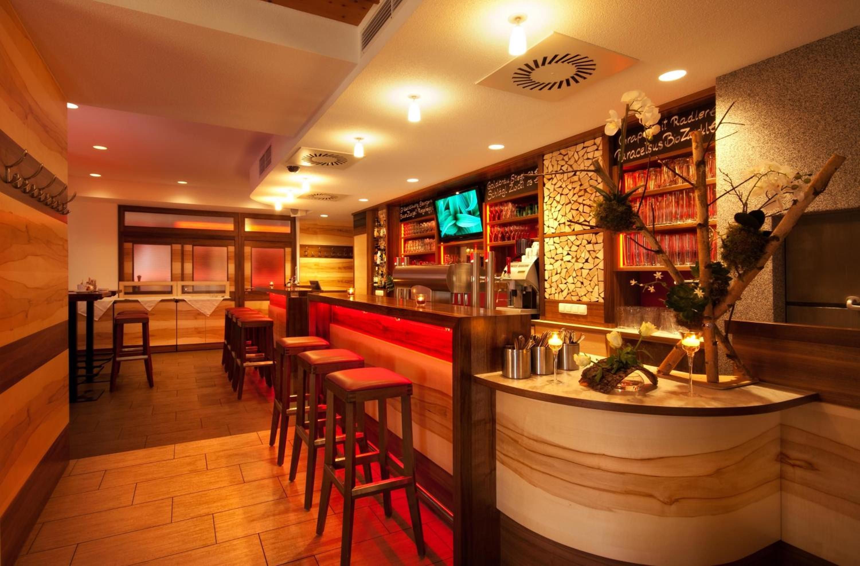 A bar area with wooden flooring, bar stools, a counter with a television, and shelves with bottles and glasses.