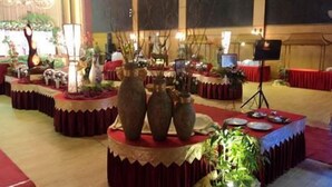 Salão de banquetes com mesas redondas cobertas por toalhas vermelhas, decorado com vasos e plantas.