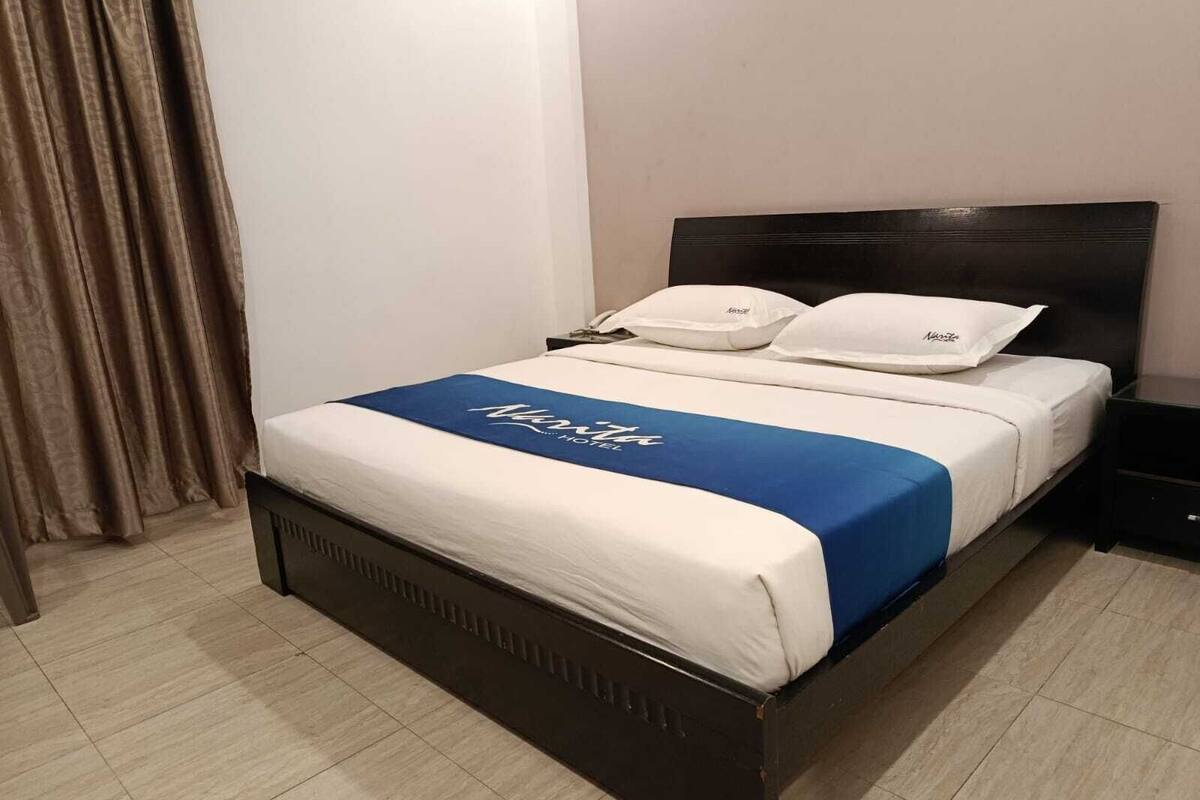 Una habitación de hotel con una cama, una mesita de noche, una colcha azul y blanca y una cortina.