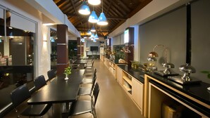 Uma sala de jantar moderna com mesas e cadeiras compridas, iluminação pendente e uma bancada de cozinha com eletrodomésticos e objetos decorativos.