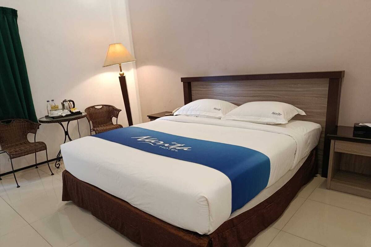 Habitación de hotel con cama, mesita de noche, lámpara, silla y pequeña mesa con una botella y vasos.