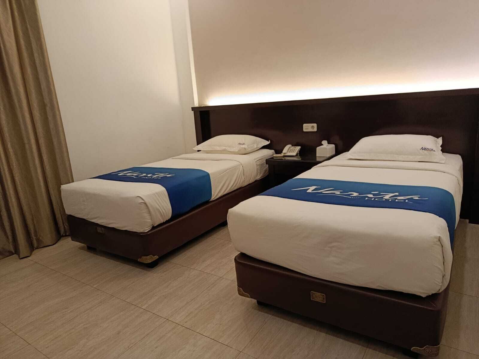 Two twin beds with white bedding and blue headboards, a bedside table with a phone, and a wall-mounted light.