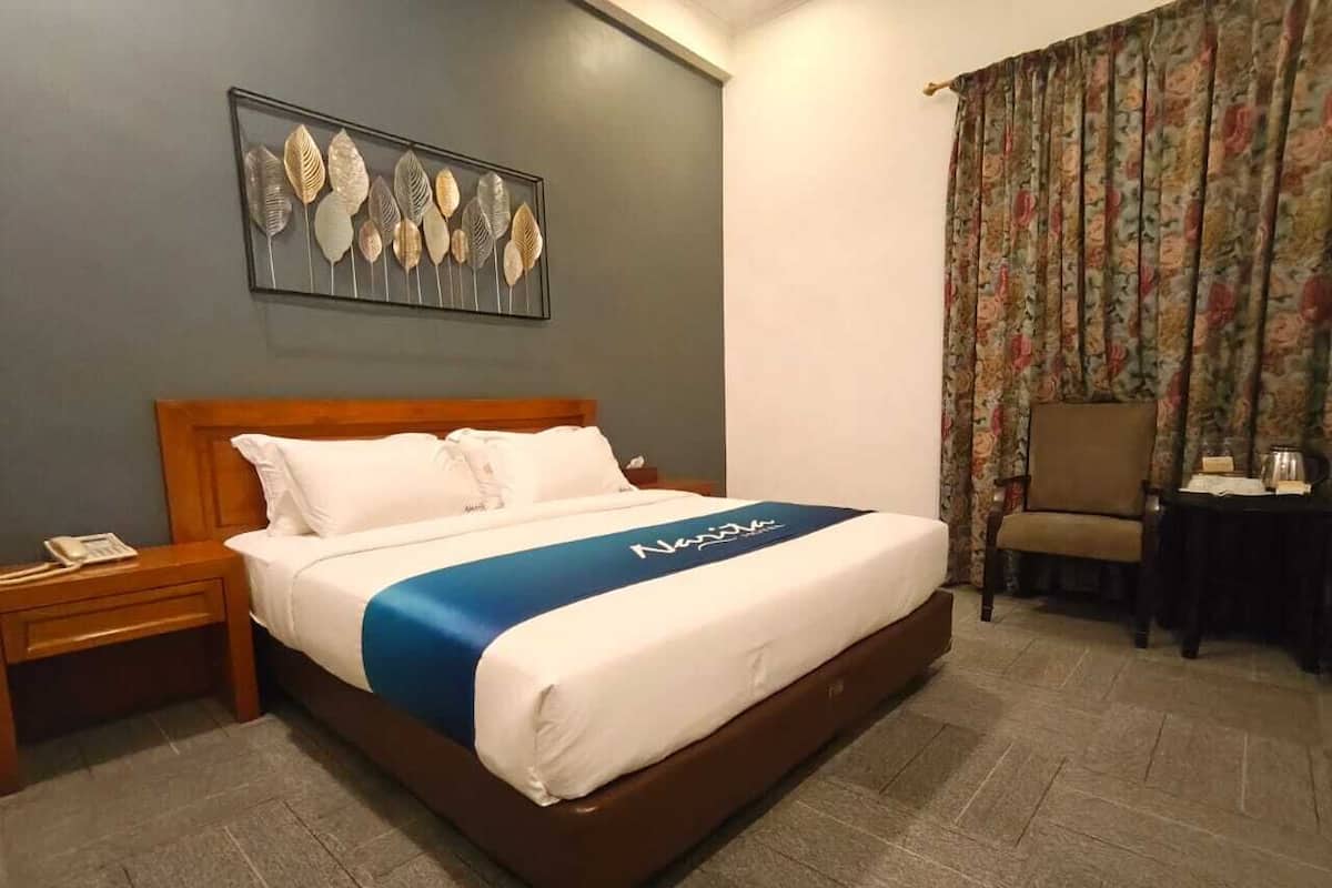 Habitación de hotel con cama, mesita de noche, silla, mesa, cortina con estampado floral y una obra de arte en la pared.