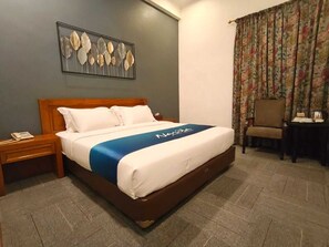 A hotel room with a bed, a nightstand, a chair, a table, a floral curtain, and a wall art piece.