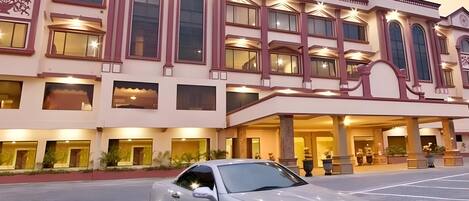 A white car parked in front of a hotel with a classic architectural style, featuring multiple windows and a grand entrance.