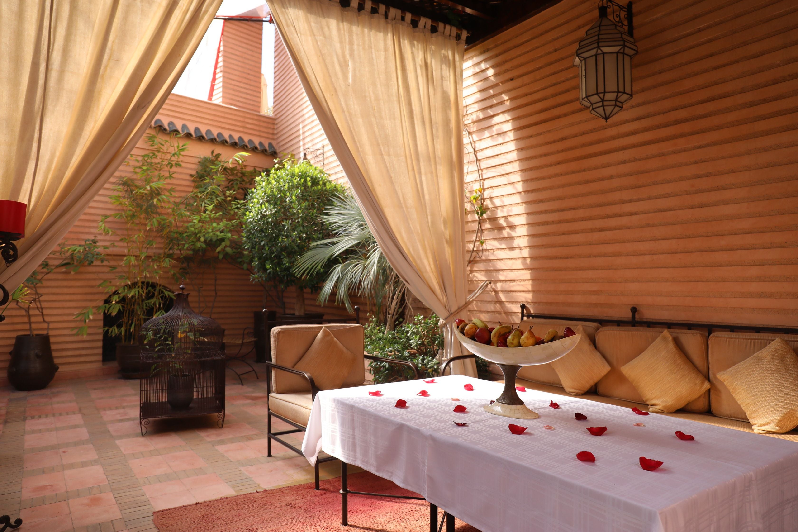 A cozy outdoor seating area with a table set for two, surrounded by beige cushions and a decorative bowl of fruit.