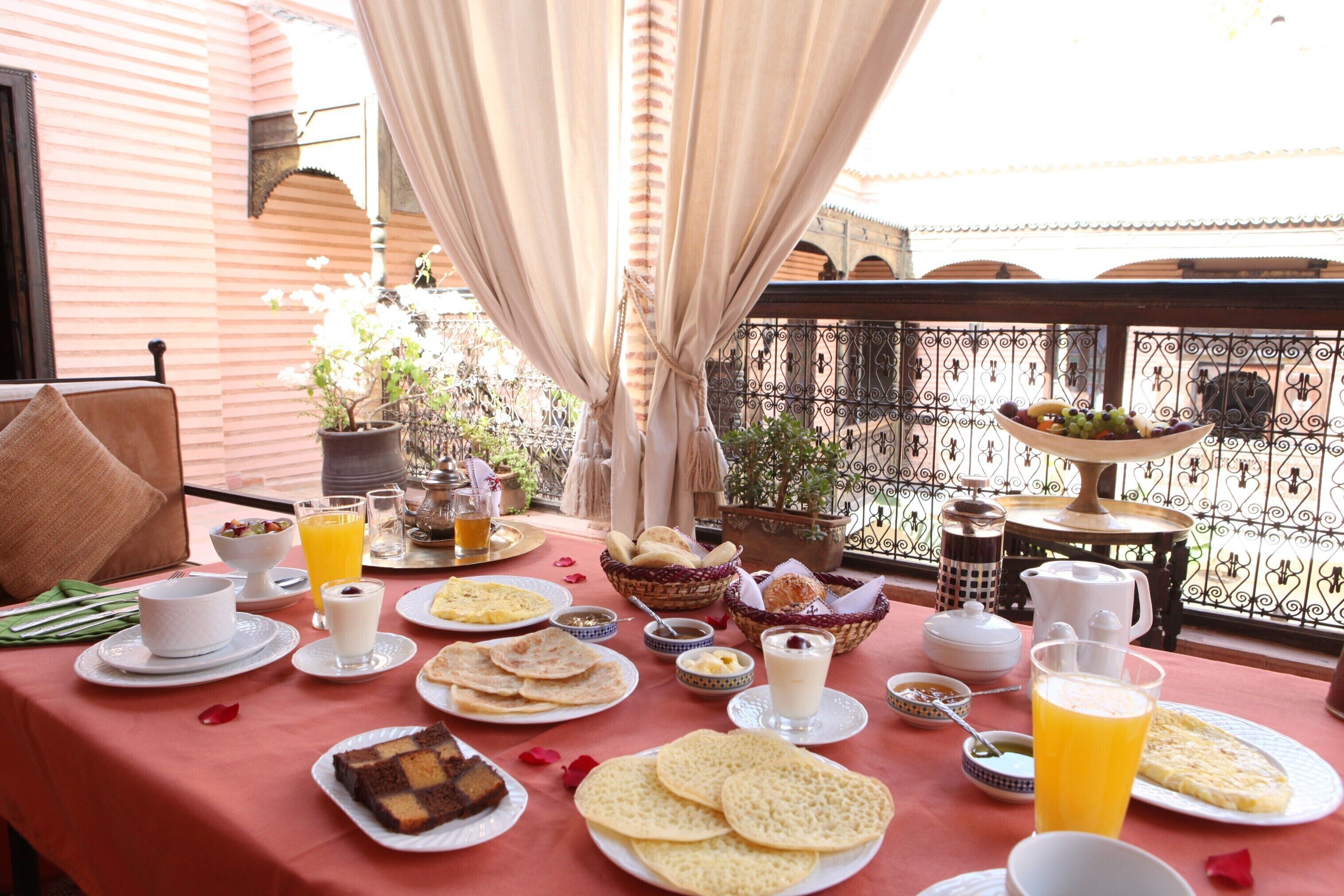 A table set with breakfast items including bread, pastries, and beverages, with a balcony view.