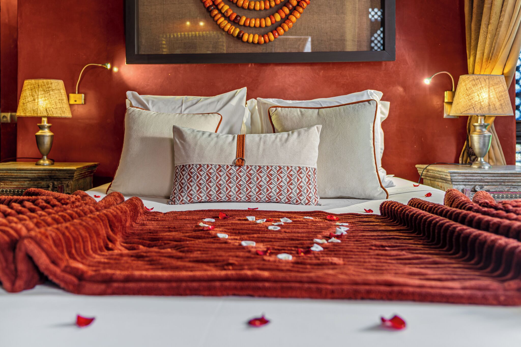 Un lit avec deux oreillers, une couverture rouge et des pétales de rose sur un couvre-lit blanc.