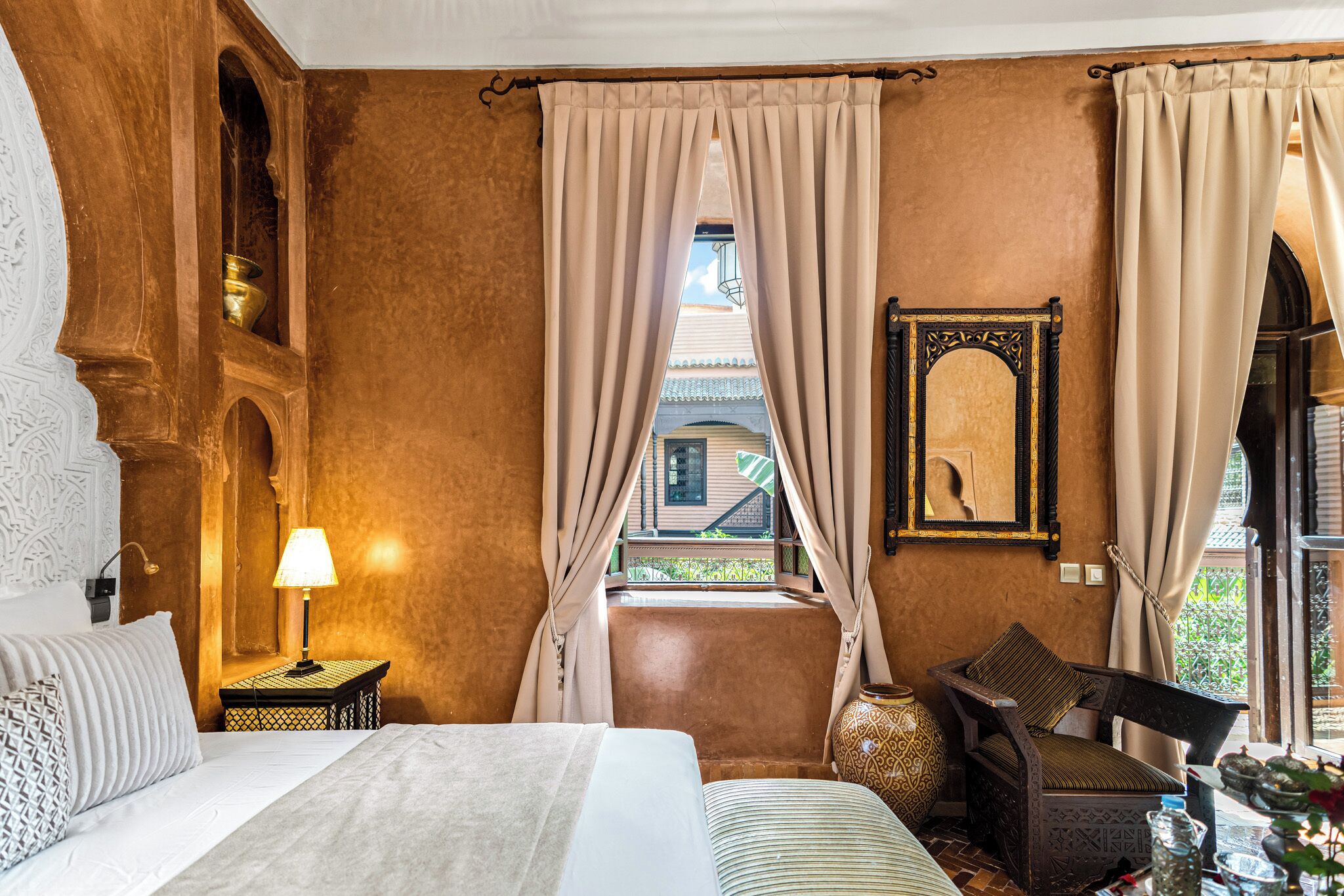 A bedroom with a bed, a nightstand with a lamp, a chair, a large vase, and a window with curtains.
